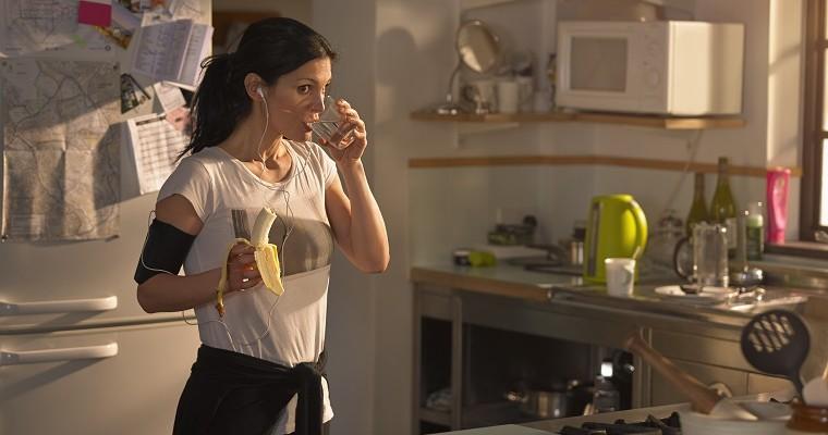 Kobieta w kuchni - pije wodę i je banana, przygotowuje się do ćwiczeń. 
