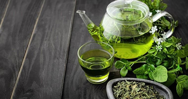 Zielona herbata zaparzona w szklanym dzbanku obok leża liście zielonej herbaty. 