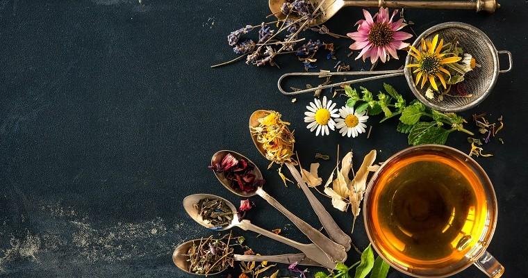 herbata ziołowa w szklance obok niej leża łyżeczki na których są rożne sypkie zioła.   