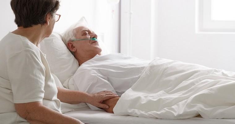 Starsza kobieta siedzi przy łóżku szpitalnym starszego mężczyzny w śpiączce